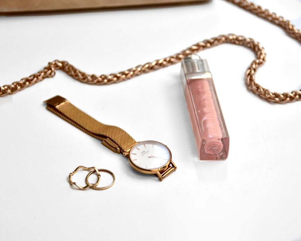 Flat lay of gold wristwatch, rings, chain, and perfume on a white surface.